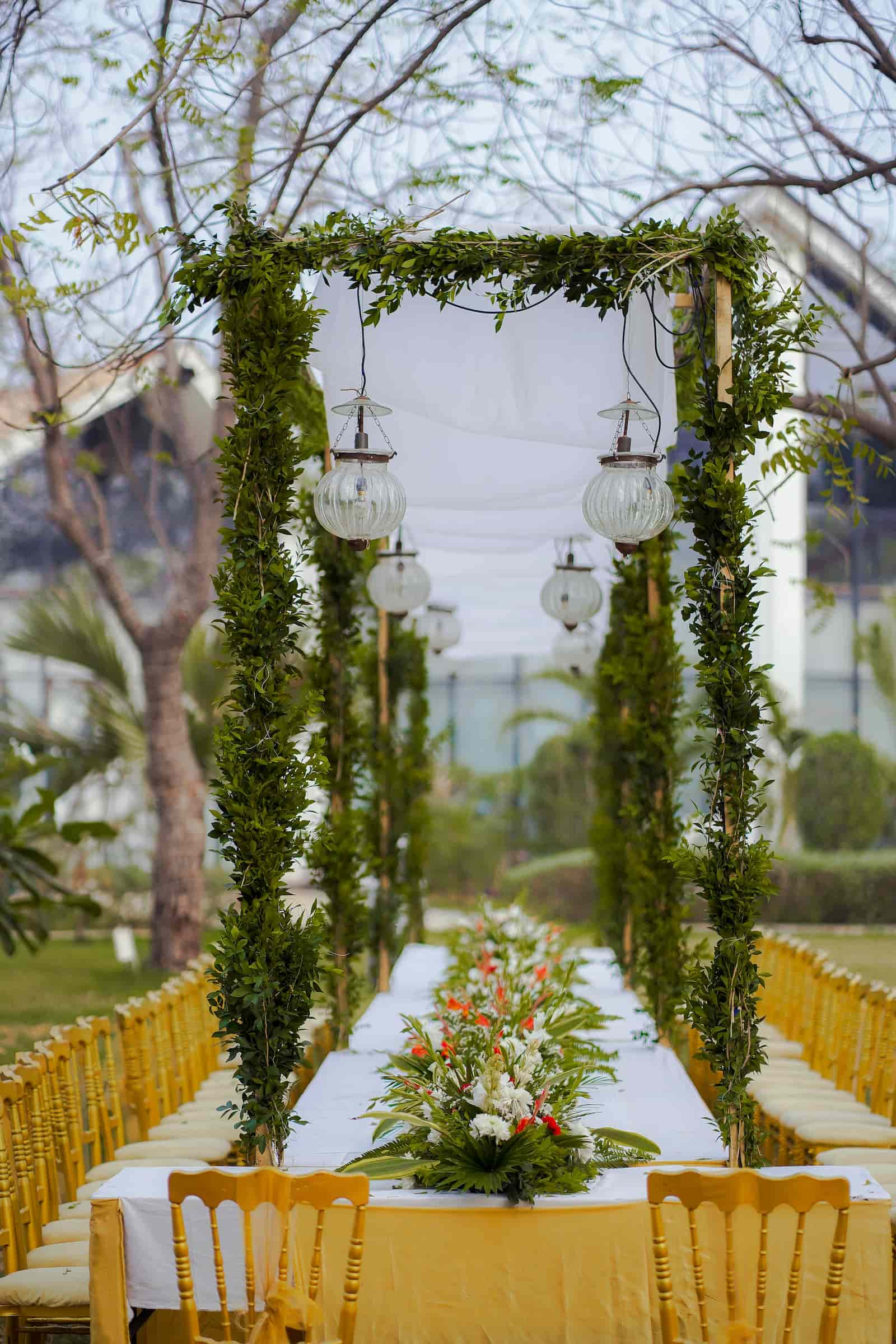 Outdoor wedding ceremony with a greenery-draped arch, hanging lanterns, and rows of gold chairs