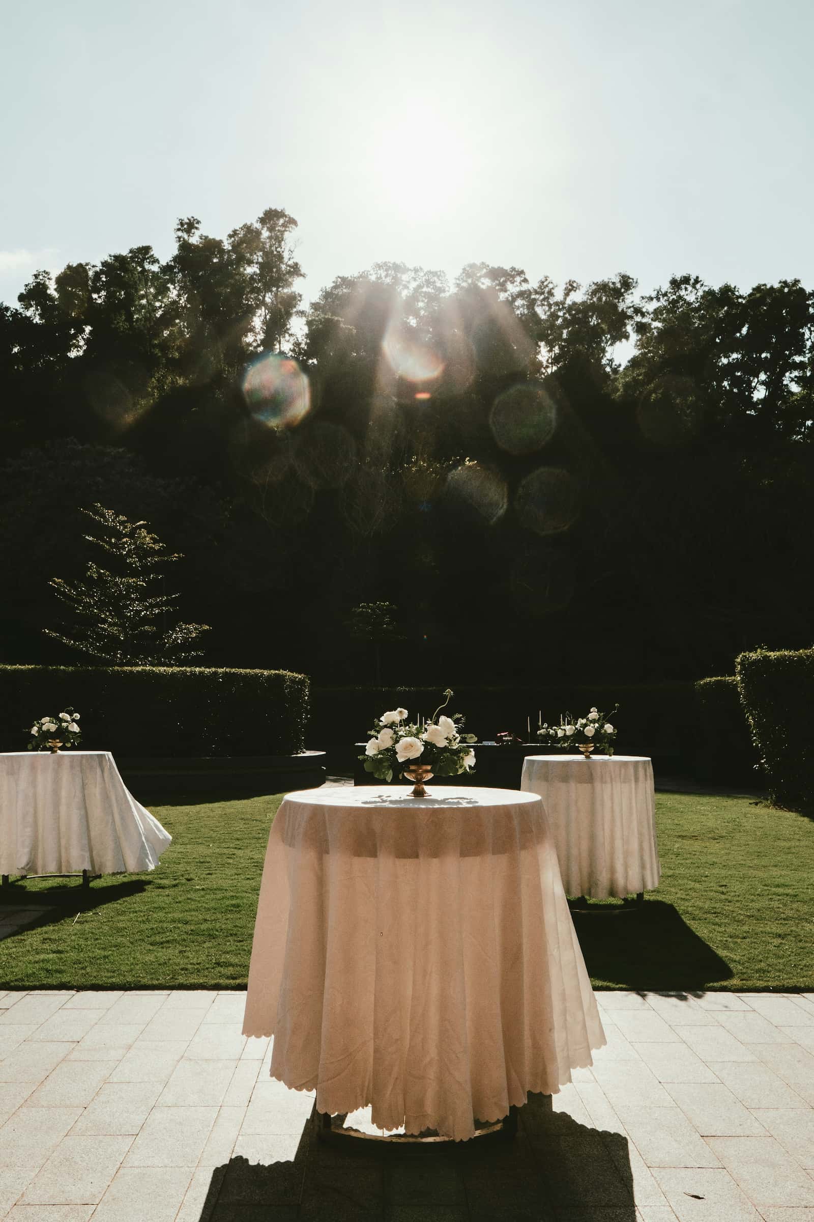 Elegant garden cocktail-hour tables with white floral centerpieces in warm afternoon light at a wedding